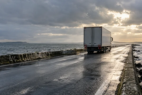 A plain pale-grey box truck sits on a wet coastal causeway, with open water on both sides and sunlight breaking through heavy clouds over the road.