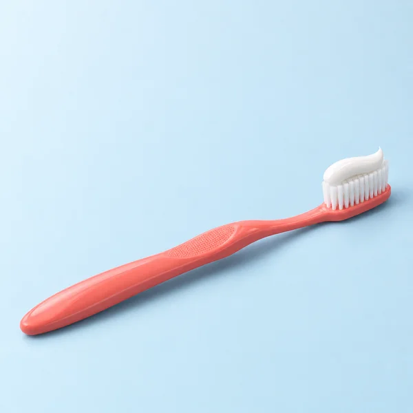 A coral manual toothbrush lies diagonally on a pale ice-blue surface, with white bristles and a curved ribbon of white toothpaste on its head.