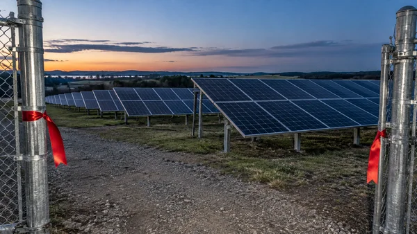 Rows of blue-black solar panels extend across a gently sloping field at dusk, viewed through a galvanized-steel gate opening.