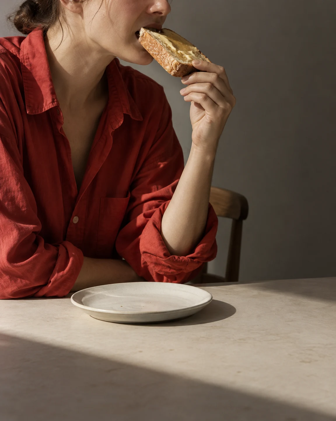 A seated adult in a loose tomato-red shirt bites buttered toast while holding it above an off-white plate on a pale table, against a warm-grey wall.