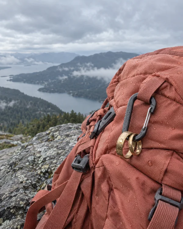 A rain-speckled coral hiking backpack fills the right side of a rocky overlook, with gold bands hanging from a large dark carabiner; a cloudy sky, distant…