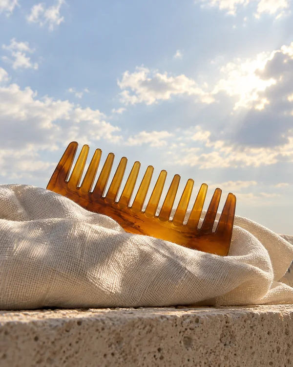 A translucent amber wide-tooth comb rests partly tucked into a loosely folded cream towel on a pale stone ledge, with its teeth upright against a pale blue sky…