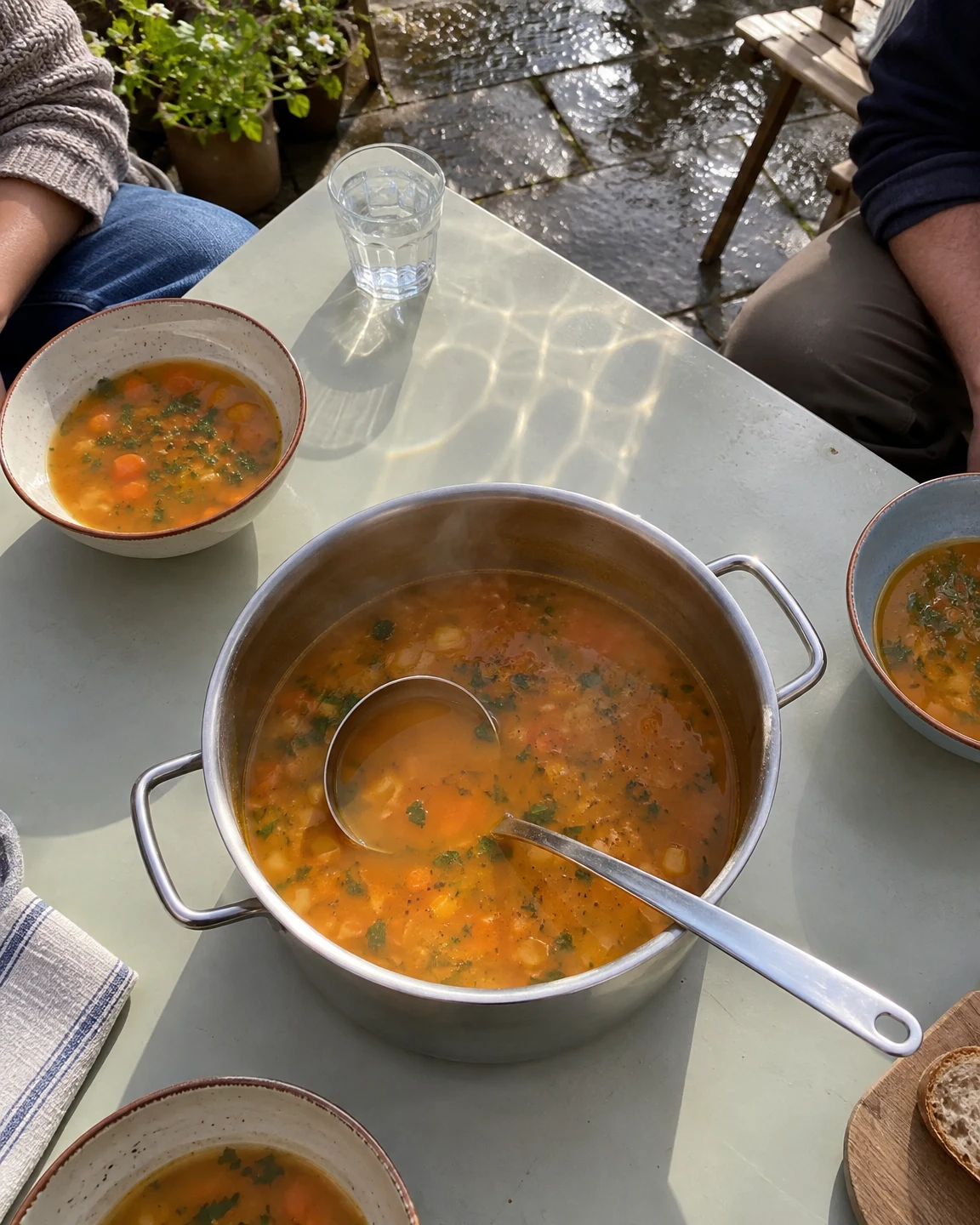 A stainless-steel pot of orange vegetable soup with a ladle sits on a pale courtyard table, surrounded by three ceramic bowls and a water glass.