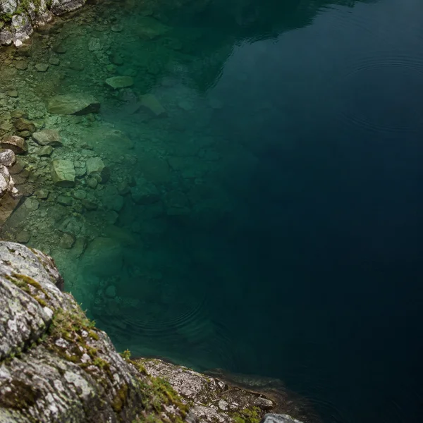 A steep view down over a rocky lakeshore, with pale submerged stones in green shallows fading into dark teal water and faint ripples.