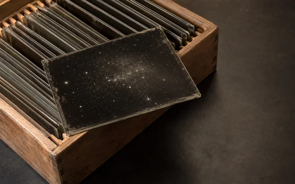 An open, worn wooden box of glass plates sits on a dark tabletop, with one star-speckled plate laid diagonally across the box edge and empty space to the right.