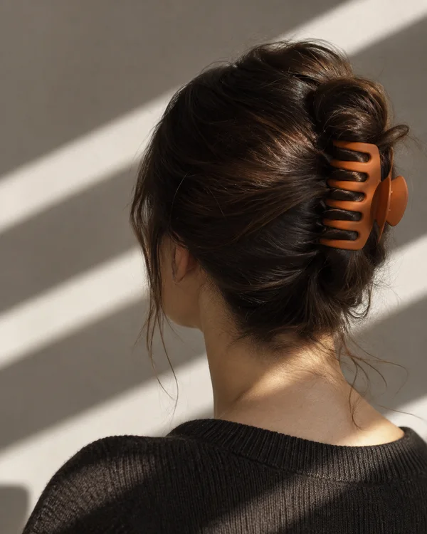 Rear three-quarter view of an adult with dark brown hair twisted up in a burnt-orange claw clip, with loose strands at the nape and a dark knit top against a…