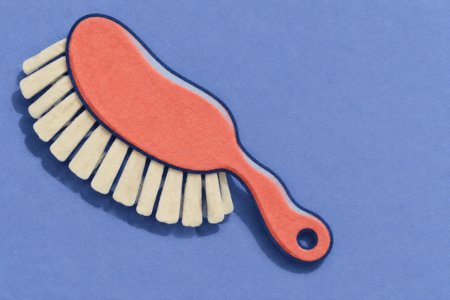 An overhead felt-style illustration of a coral brush with oversized cream bristle tabs and a rounded hanging hole, lying diagonally on a periwinkle-blue…