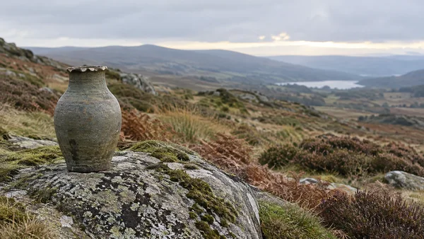 A weathered olive-grey stoneware vase with a chipped rim stands on a lichen-covered granite boulder in the foreground of a quiet upland moor, with heather…