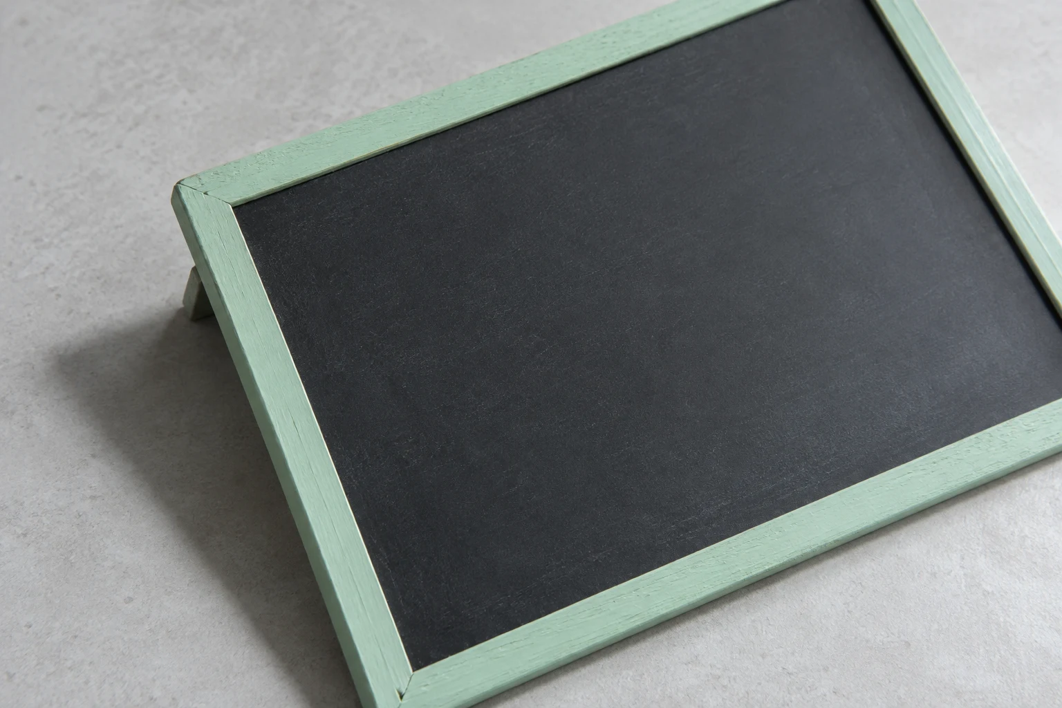 A blank charcoal chalkboard in a mint-painted wooden frame lies diagonally on a pale grey tabletop, with its folding support and a soft shadow visible beneath…