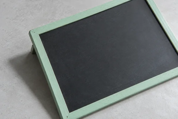 A blank charcoal chalkboard in a mint-painted wooden frame lies diagonally on a pale grey tabletop, with its folding support and a soft shadow visible beneath…