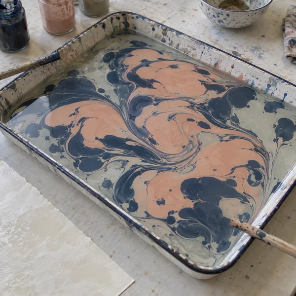 A rectangular enamel tray of peach and navy marbling ink sits diagonally on a pale workshop table, with stirring sticks at opposite edges and a blank sheet of…
