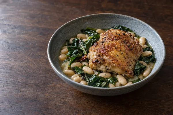 A roasted, skin-on chicken thigh sits over white beans and wilted greens in a blue-gray ceramic bowl on a dark walnut tabletop, with open space to the left.