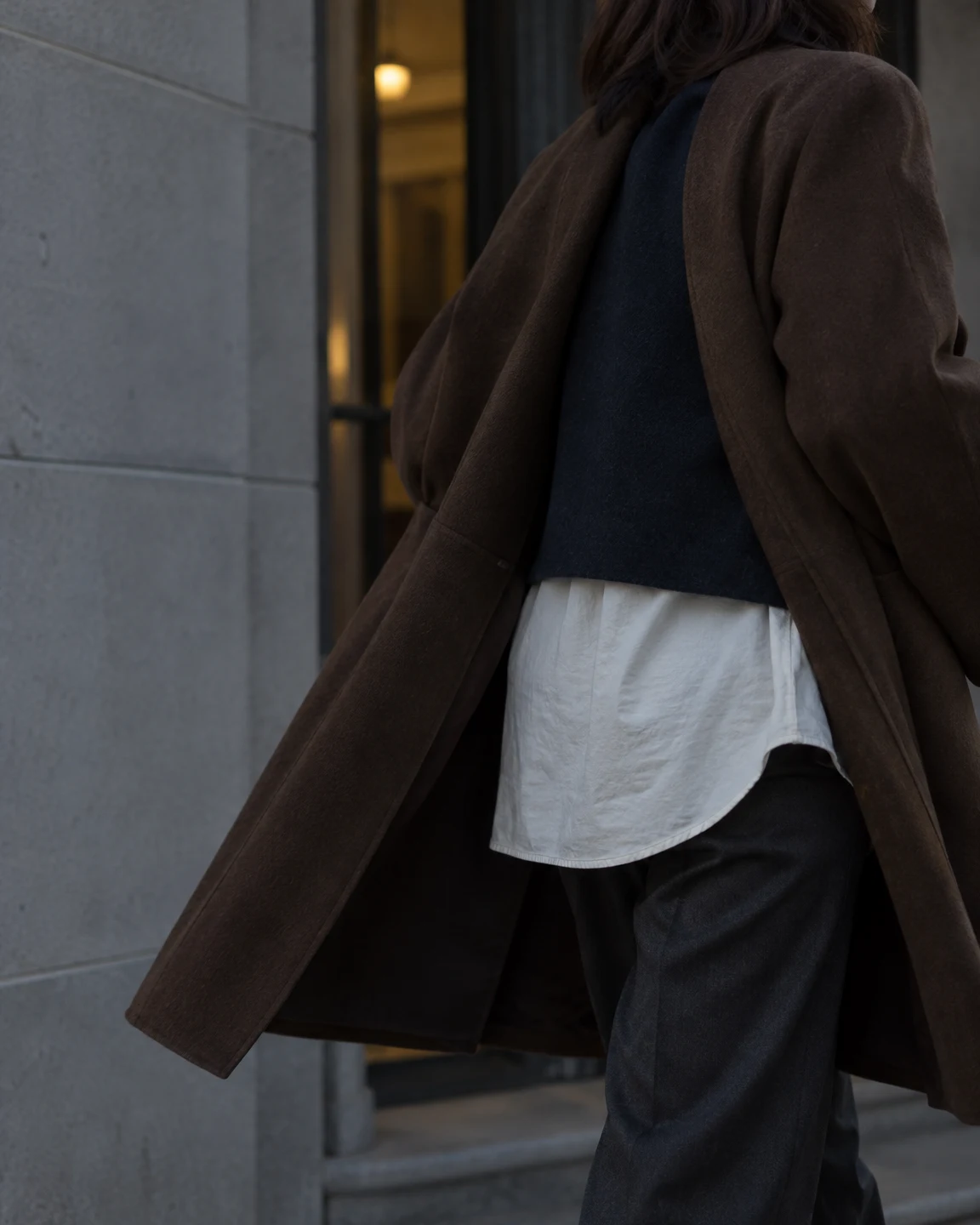 A person in an open tobacco-brown coat, navy vest, ivory shirt, and dark trousers passes a recessed entrance beside a grey stone wall, with warm light visible…