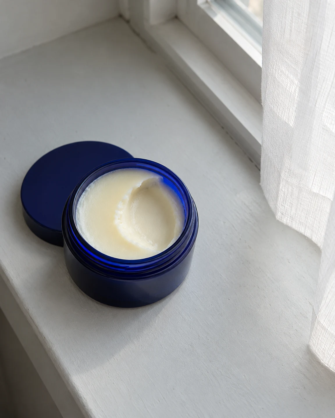 An open cobalt-blue glass jar of ivory balm with a broad curved scoop sits on a pale painted windowsill, with its lid partly beneath it and a sheer curtain…