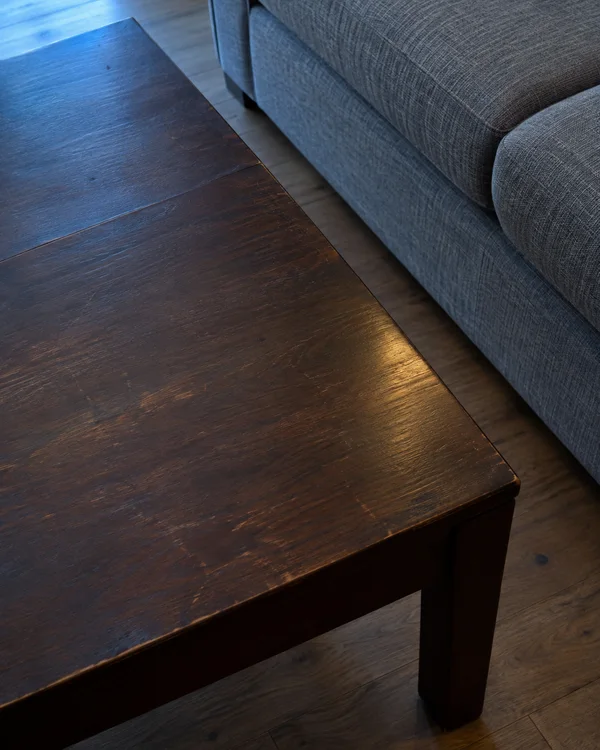 Oblique overhead view of a dark-brown wooden coffee table beside a grey woven sofa, with a strip of wood flooring visible between them and one table leg in…