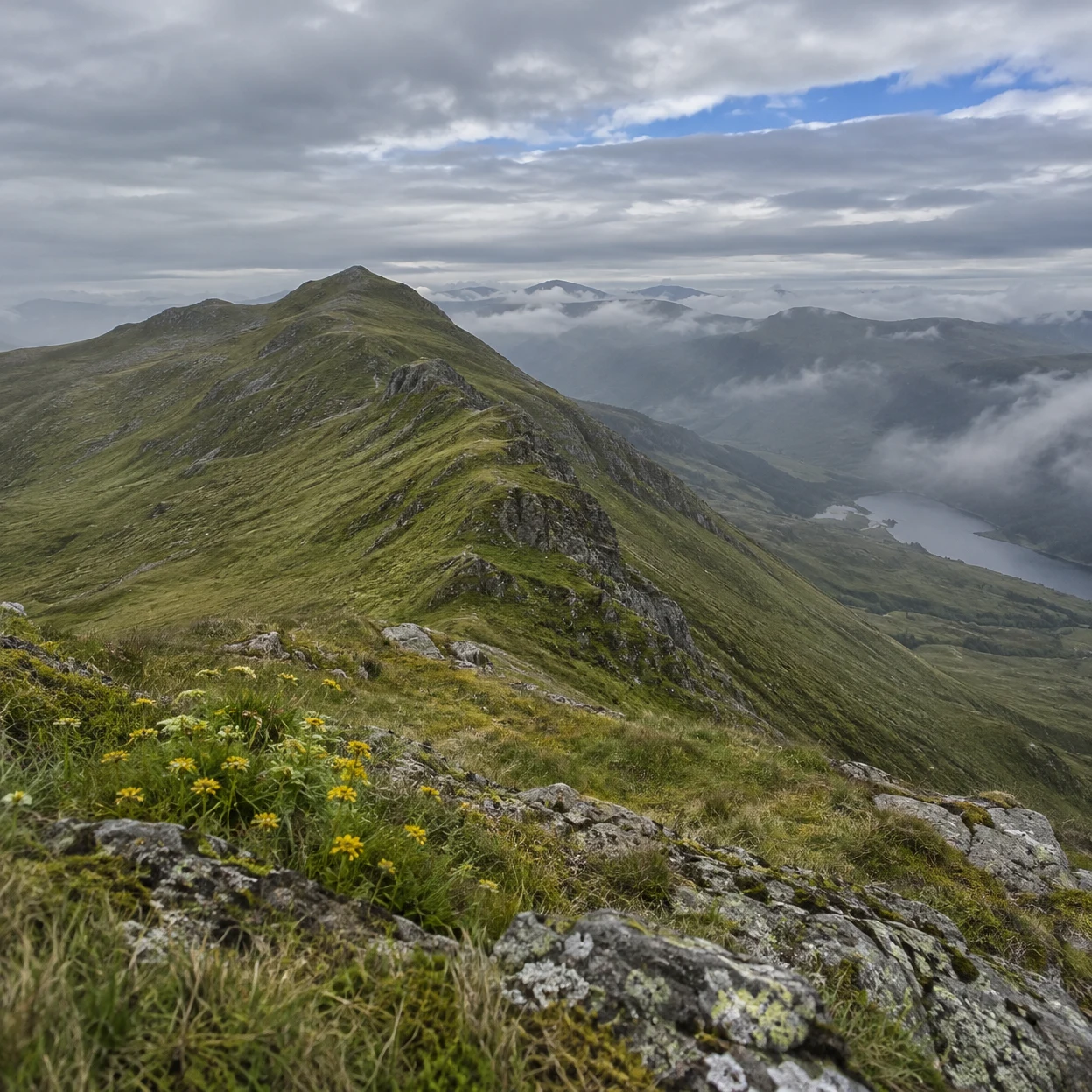 A grassy mountain ridge with rocky, lichen-covered foreground slopes rises above a misty valley under layered grey clouds, with a blue opening in the sky and…