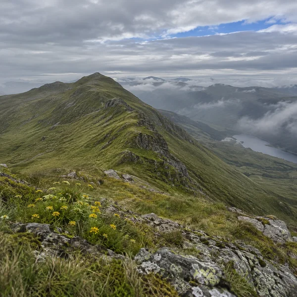 A grassy mountain ridge with rocky, lichen-covered foreground slopes rises above a misty valley under layered grey clouds, with a blue opening in the sky and…