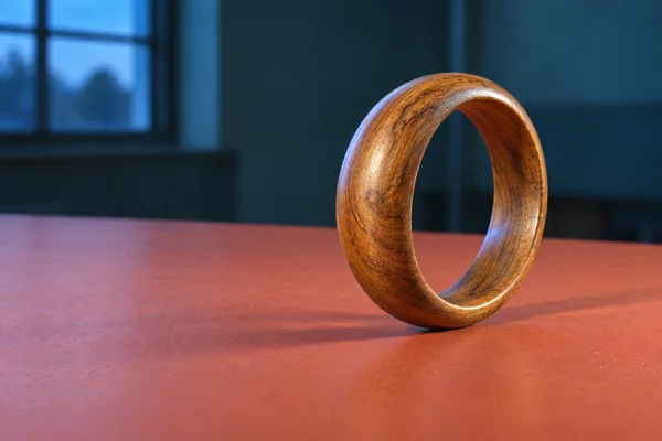 A thick honey-brown wooden bangle stands on its lower rim on a coral workbench, with its circular opening visible against a dark teal interior and a blue-lit…