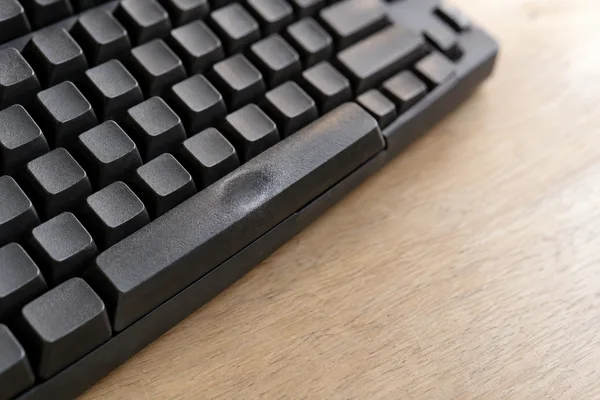 A close, oblique view of a charcoal keyboard on a pale wooden desk, with a shiny worn patch on the spacebar and empty desktop to the right.