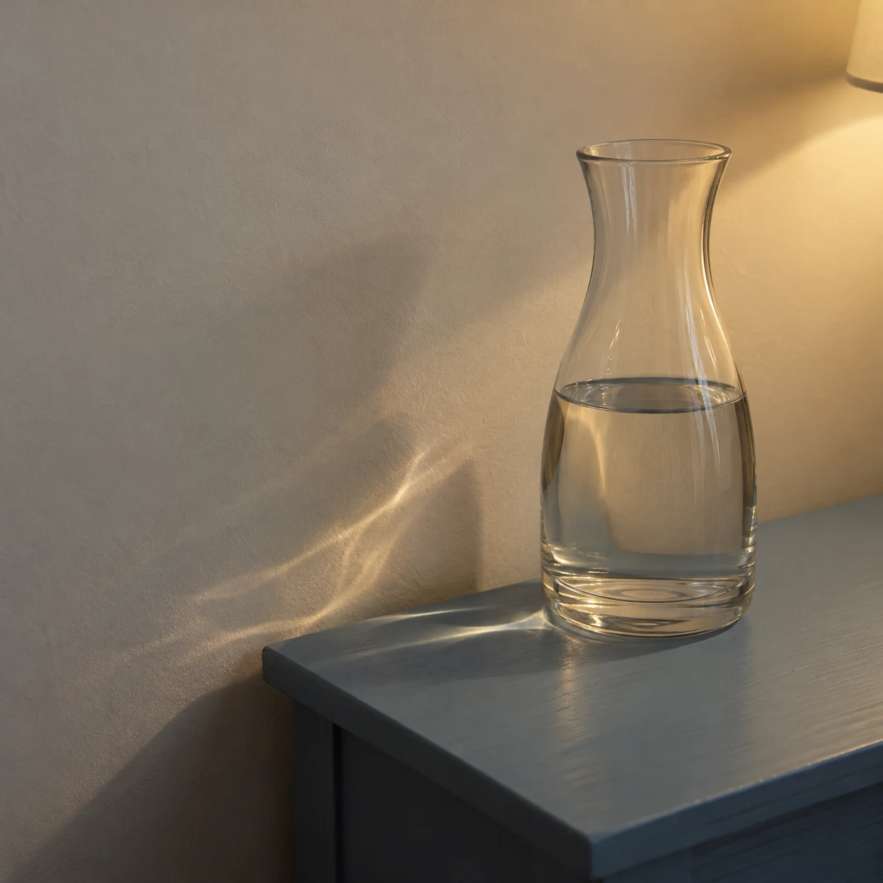 A clear glass carafe, about half full of water, sits right of center on a dark blue ledge against a warm off-white wall.