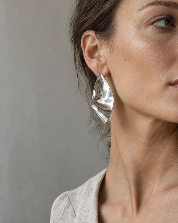 A close side crop of a woman with dark hair tucked behind her ear, wearing a large reflective sculptural silver earring against a plain warm-grey wall; part of…