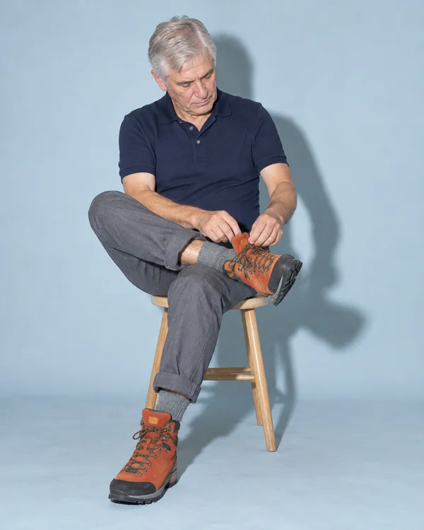 A silver-haired older man in a navy polo and cuffed gray trousers sits cross-legged on a wooden stool against a pale blue backdrop, looking down as he adjusts…