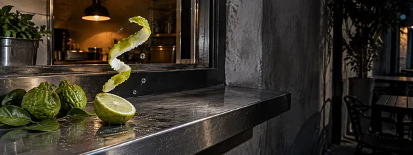 A curled yellow-green citrus peel hangs above a cut citrus half and several whole bumpy green fruits on a reflective steel café counter.