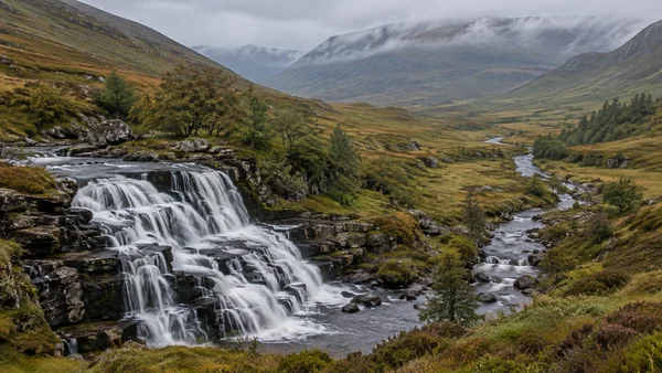 A broad stepped waterfall pours over dark rock ledges in the lower left of a grassy upland valley, with a winding river leading toward mist-covered hills…