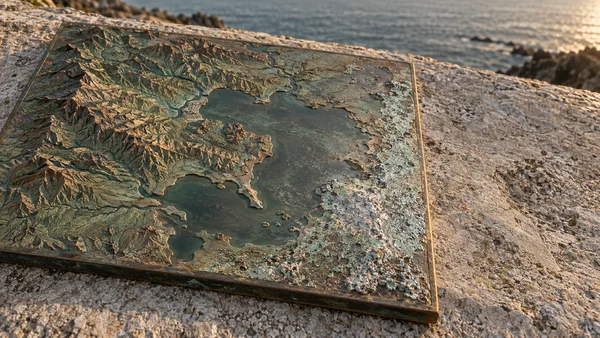 A rectangular copper relief map with raised, rugged contours and blue-green patina rests on a sunlit stone ledge, with a blurred strip of sea beyond it.