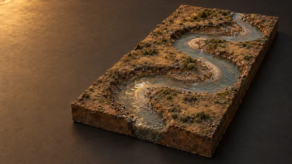 A rectangular miniature terrain slab sits on a dark tabletop, with a shallow winding river channel cutting through rough ochre ground and catching warm…