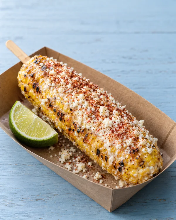 A grilled corn cob topped with creamy white cheese, browned grill marks and red chile seasoning rests diagonally in a kraft-paper tray on a pale-blue wooden…