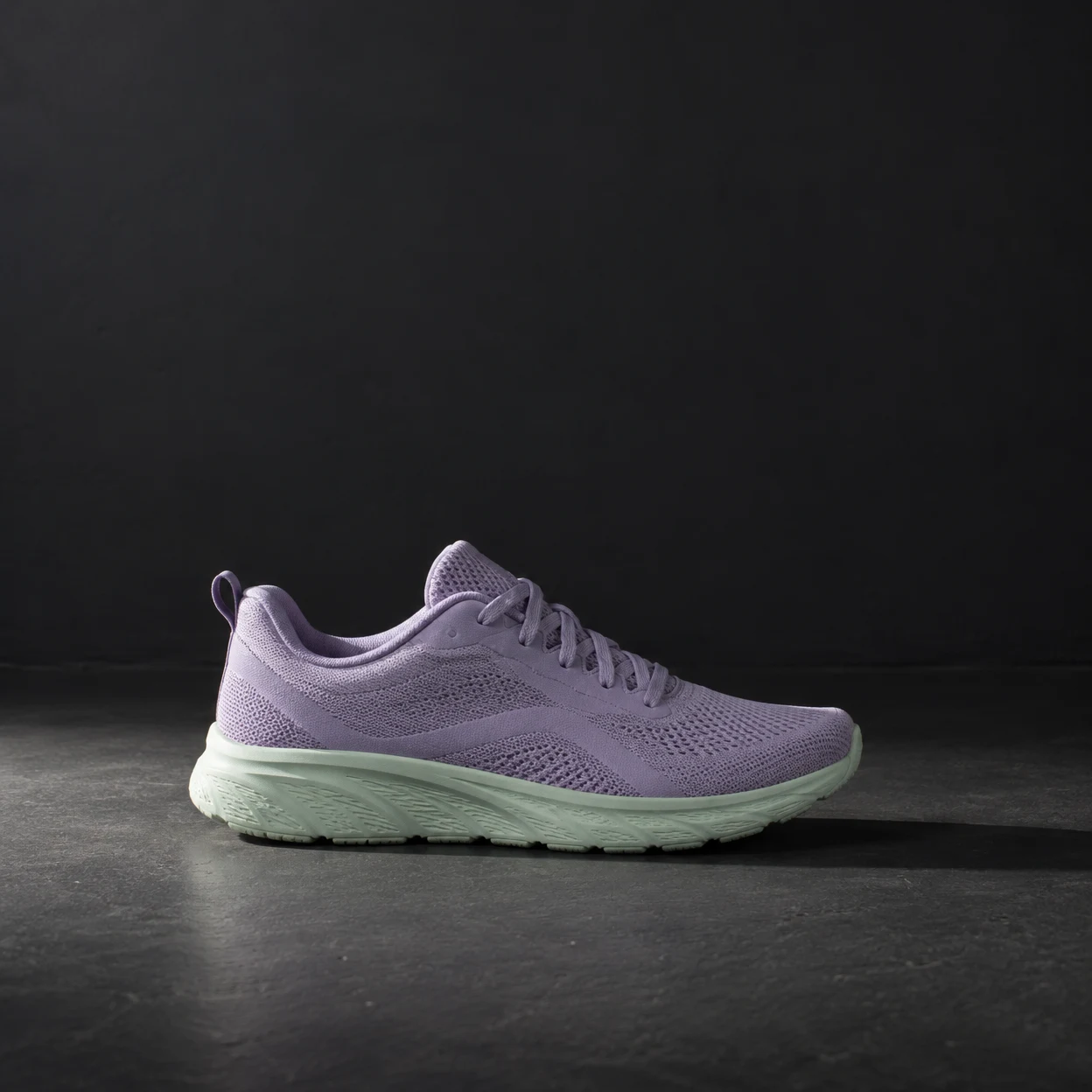 A lavender knit running shoe with a pale mint sole rests in lateral profile on a dark stage floor, its toe pointing right beneath a plain charcoal wall.
