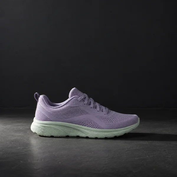 A lavender knit running shoe with a pale mint sole rests in lateral profile on a dark stage floor, its toe pointing right beneath a plain charcoal wall.