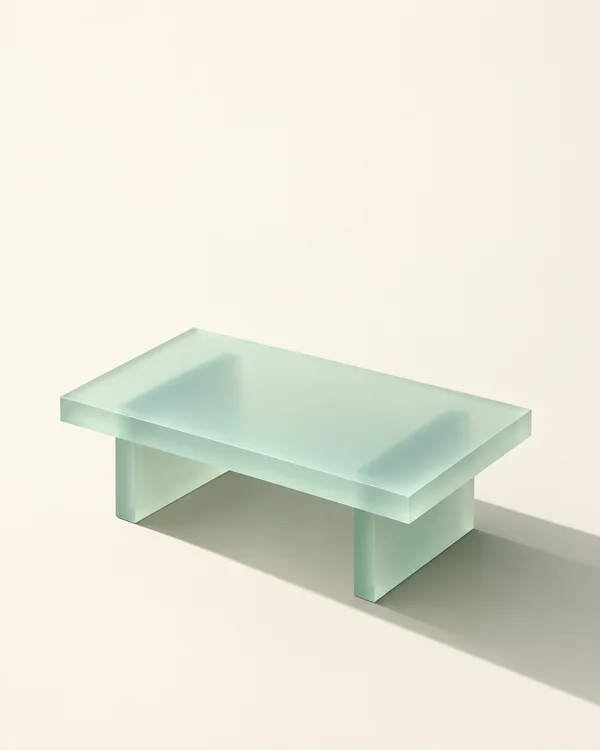 A low mint-green translucent coffee table with a thick rectangular top and two broad slab supports sits on a warm ivory ground, casting a long shadow toward…