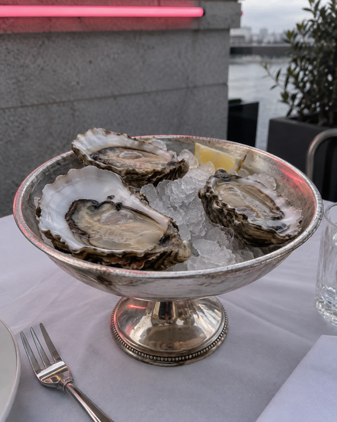 Three opened oysters rest on crushed ice in a footed silver serving dish on white table linen.