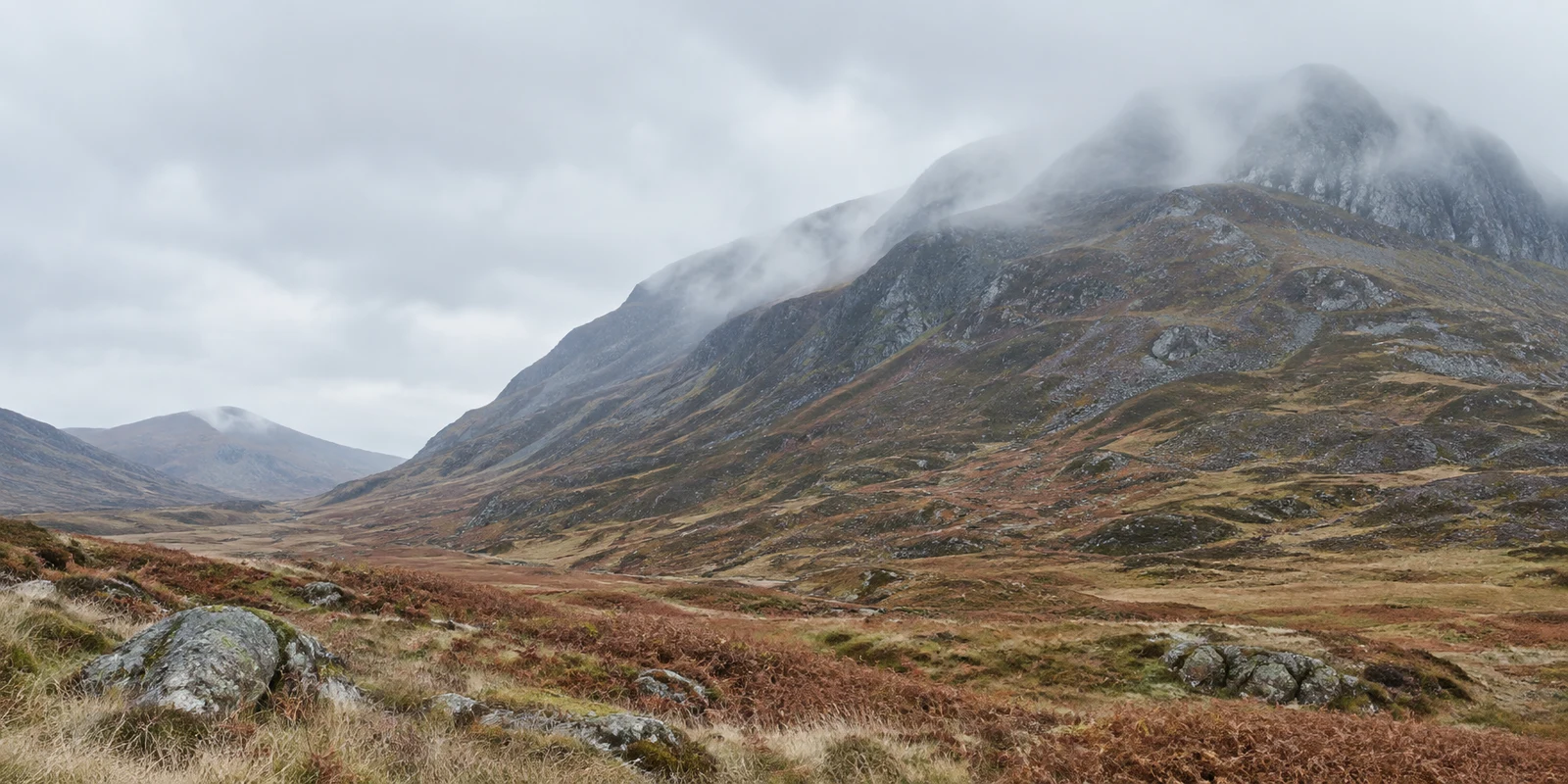 A broad rocky mountain rises above a rust-brown upland valley, with low mist drifting across its upper slopes beneath a pale grey sky.