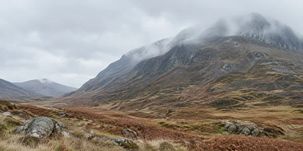 A broad rocky mountain rises above a rust-brown upland valley, with low mist drifting across its upper slopes beneath a pale grey sky.