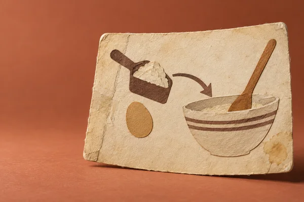 A large worn cream recipe card stands against a dusty terracotta background, with layered paper shapes showing a flour scoop, an egg, a curved arrow, and a…