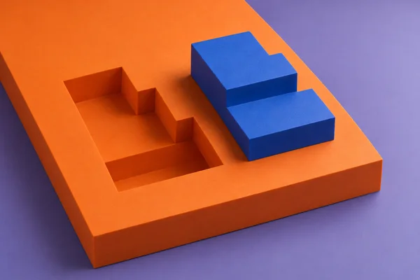 A cobalt-blue stepped block rests beside a matching stepped recess in a large orange slab on a muted violet surface, viewed from above at an angle.