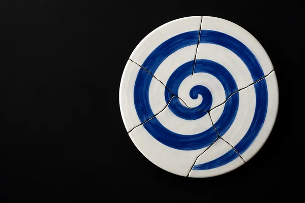 A white ceramic disc assembled from irregular fragments sits right of center on a dark surface, with a broad cobalt-blue spiral pattern crossing the visible…