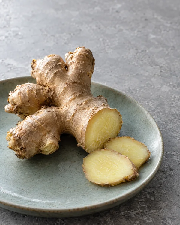A branching ginger rhizome with a cut end and two thick slices rests on a pale celadon plate on gray stone, with open space above it.