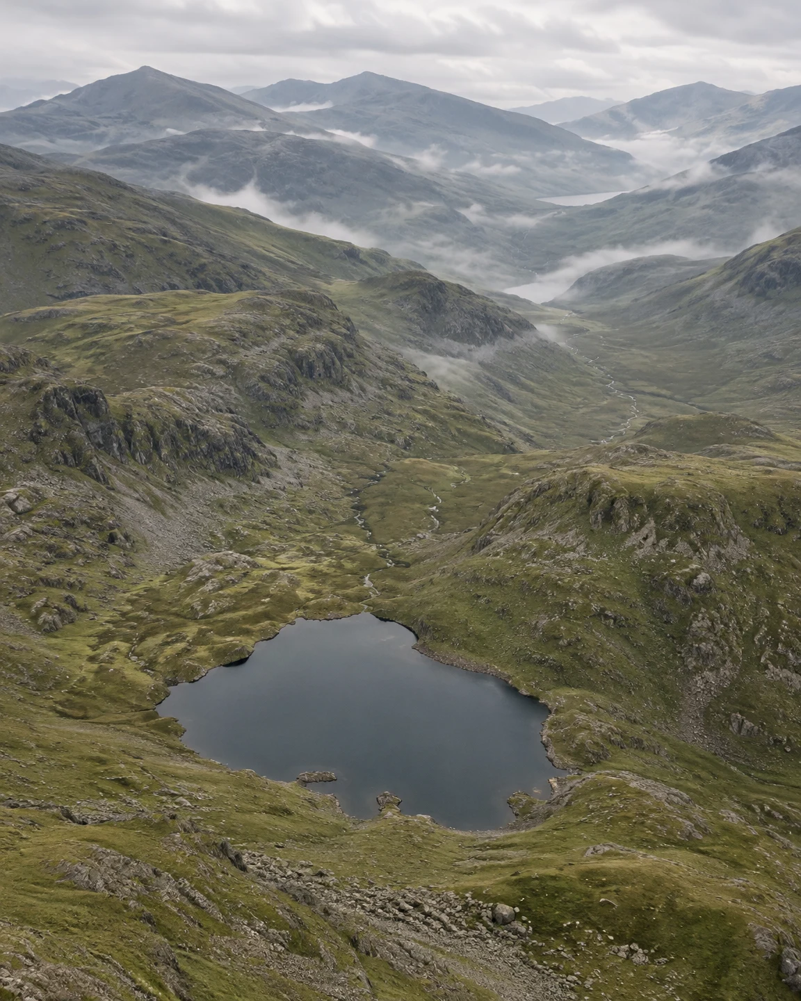 A dark mountain tarn sits in the foreground of a broad grassy valley, with a narrow stream winding between rocky slopes and mist drifting across distant…