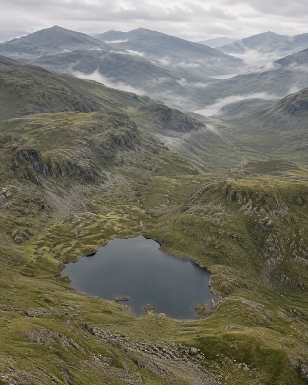 A dark mountain tarn sits in the foreground of a broad grassy valley, with a narrow stream winding between rocky slopes and mist drifting across distant…
