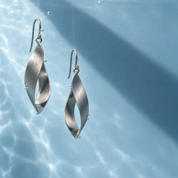A pair of satin-brushed silver-tone folded ribbon earrings hangs in clear water against an ice-blue background, with fine suspension lines, small bubbles…