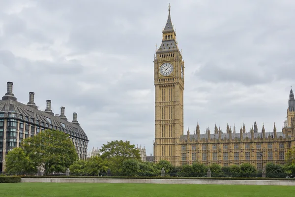 Elizabeth Tower rises above the Palace of Westminster beneath a pale overcast sky, with a lawn and stone edging in the foreground.