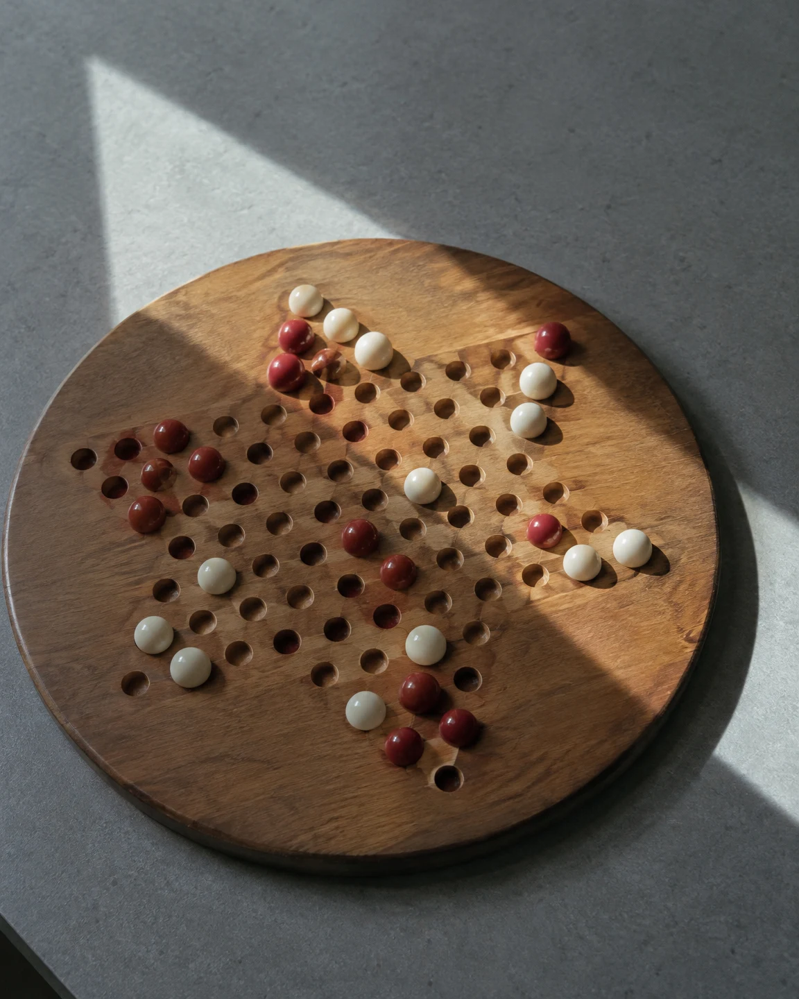A round wooden game board with rows of holes and scattered red and ivory marbles sits on a grey tabletop, crossed by large angular patches of light and shadow.