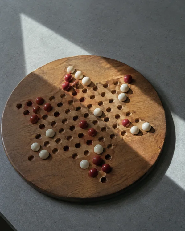 A round wooden game board with rows of holes and scattered red and ivory marbles sits on a grey tabletop, crossed by large angular patches of light and shadow.