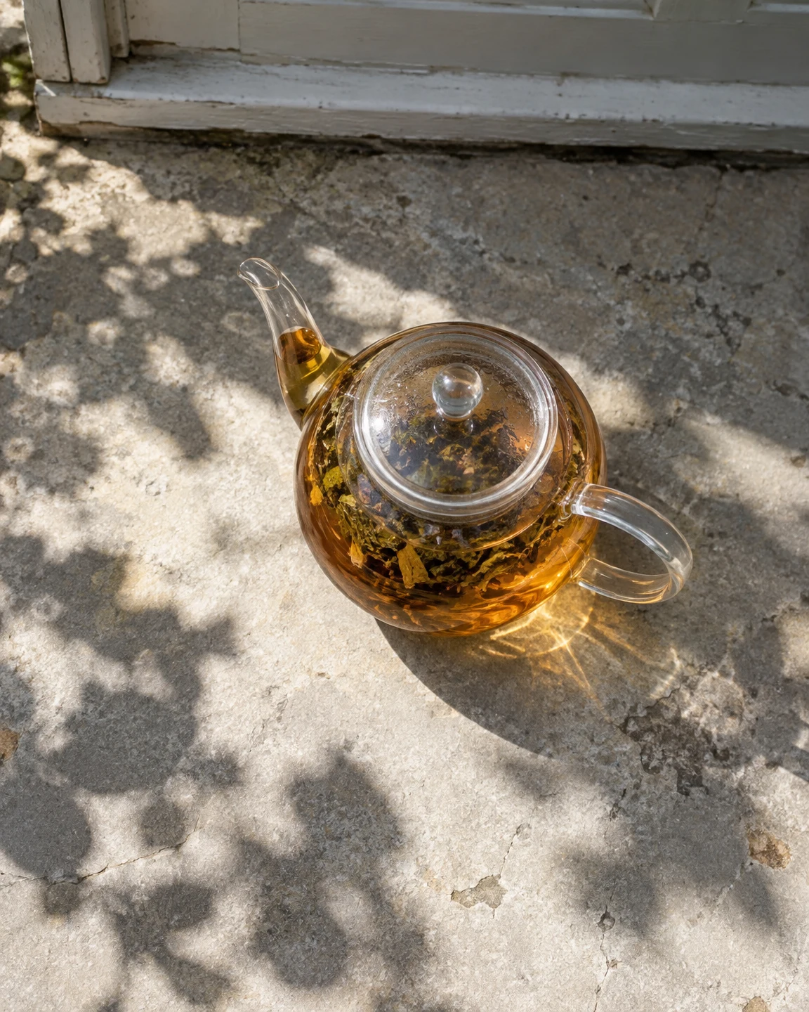 An overhead view of a clear glass teapot filled with amber tea and loose leaves, set on a sunlit, weathered stone doorstep beneath a painted sill.