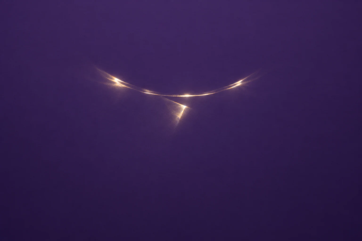 A slender warm gold-white curved light reflection and a small tilted flare sit above the centre of a deep violet field, with broad empty space around them.
