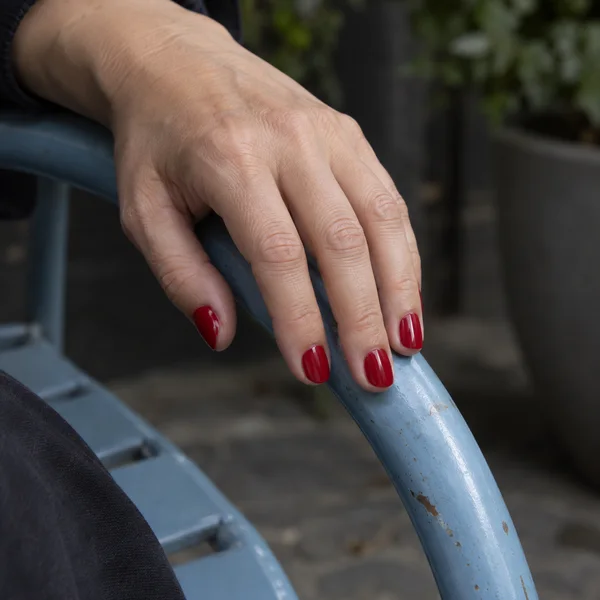 A close-up of a hand with glossy red nails resting over the curved, worn pale-blue metal arm of an outdoor chair, against a softly blurred shaded background.